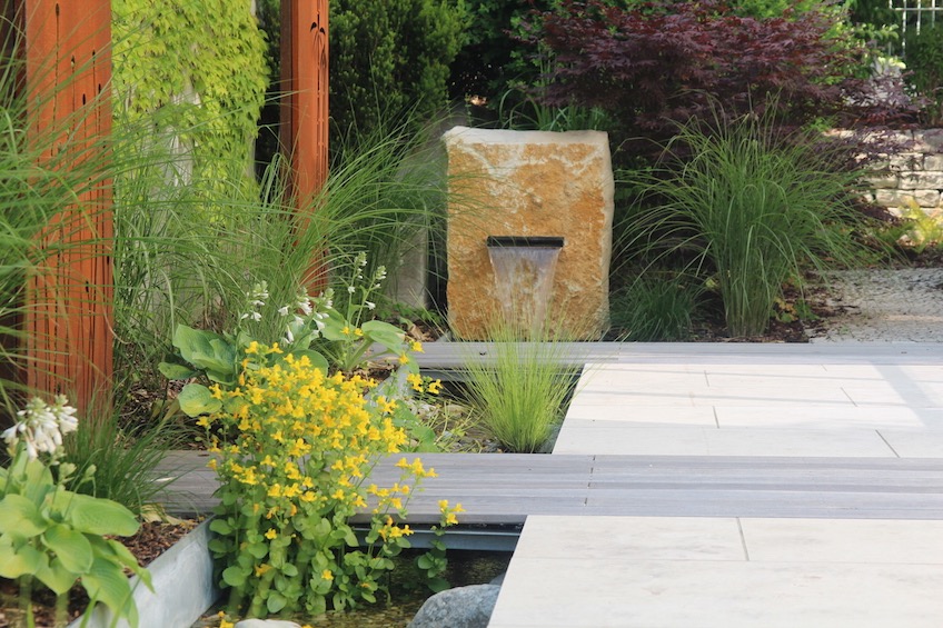 Der Bachlauf entspringt in einem Quader aus Jurakalkstein und f&uuml;hrt geradlinig am Haus entlang. Er wird von in die Steinfl&auml;che eingearbeiteten Holzstegen unterbrochen, die in Kunstw&auml;nden aus Cortenstahl enden. (Zolling bei Freising)